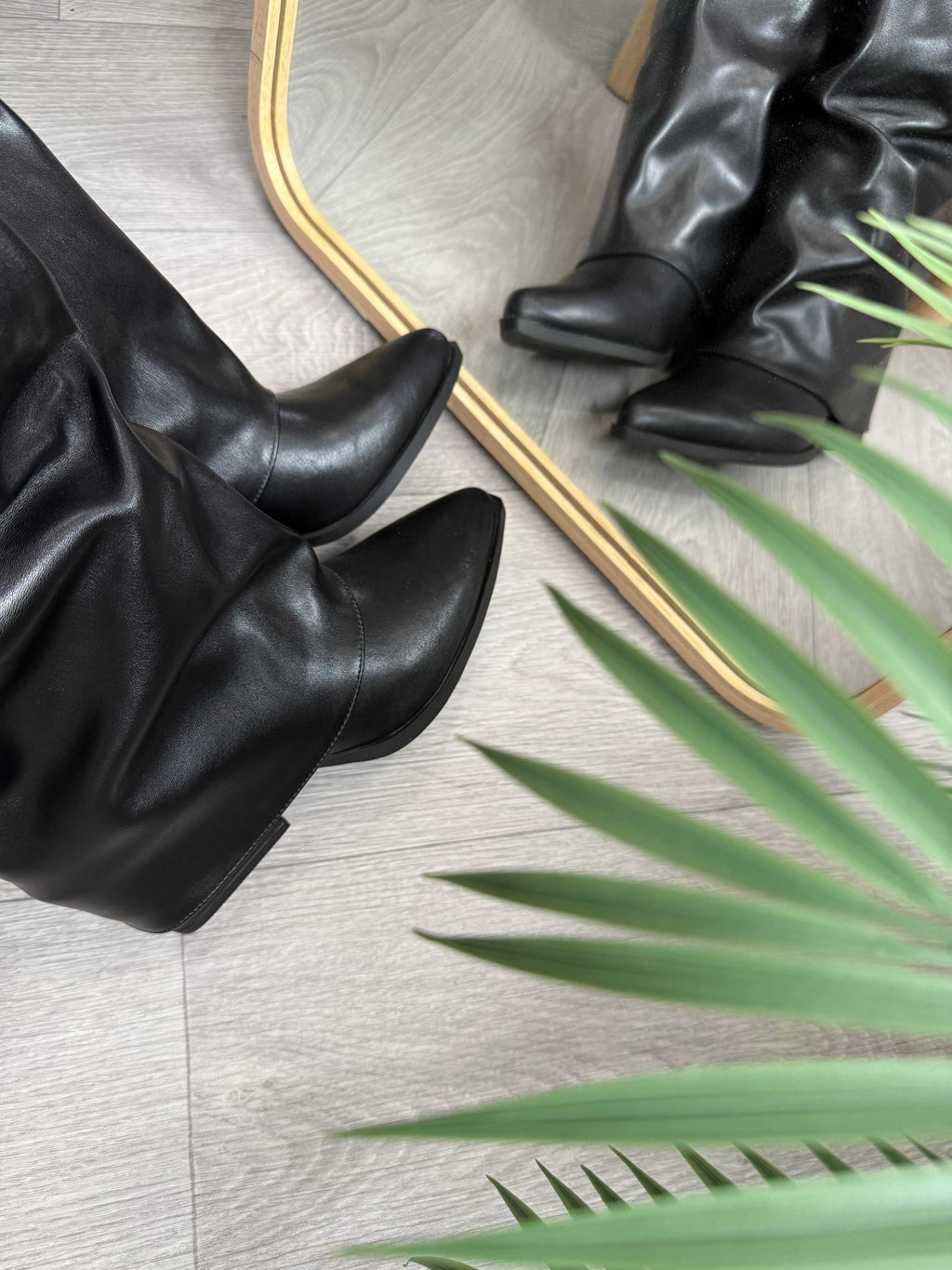 Stivale Nero black boot with maxi cuff and 5cm heel reflected in mirror on wooden floor with green plant nearby