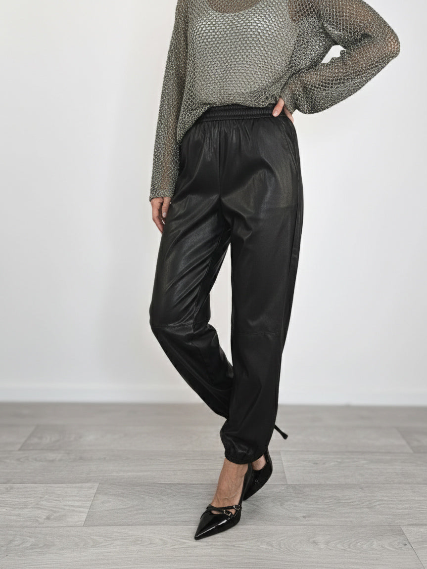 Modella indossa jogger neri in ecopelle con elastico in vita e alle caviglie, abbinati a maglia traforata grigia e tacchi a spillo neri.