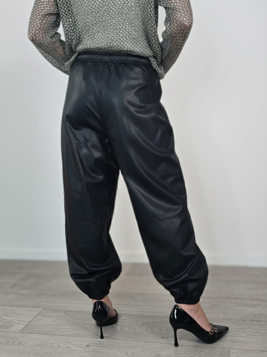 Modella indossa jogger neri in ecopelle con elastico in vita e alle caviglie, abbinati a maglia traforata grigia e tacchi a spillo neri.